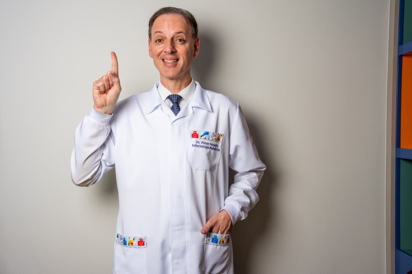 Dr. Victor Horácio sorri e aponta o dedo para cima, de jaleco no consultório